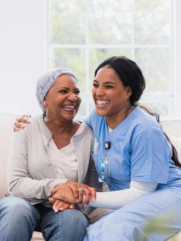 在她访问期间, the mid adult home health nurse and the senior adult woman with cancer embrace and laugh together.